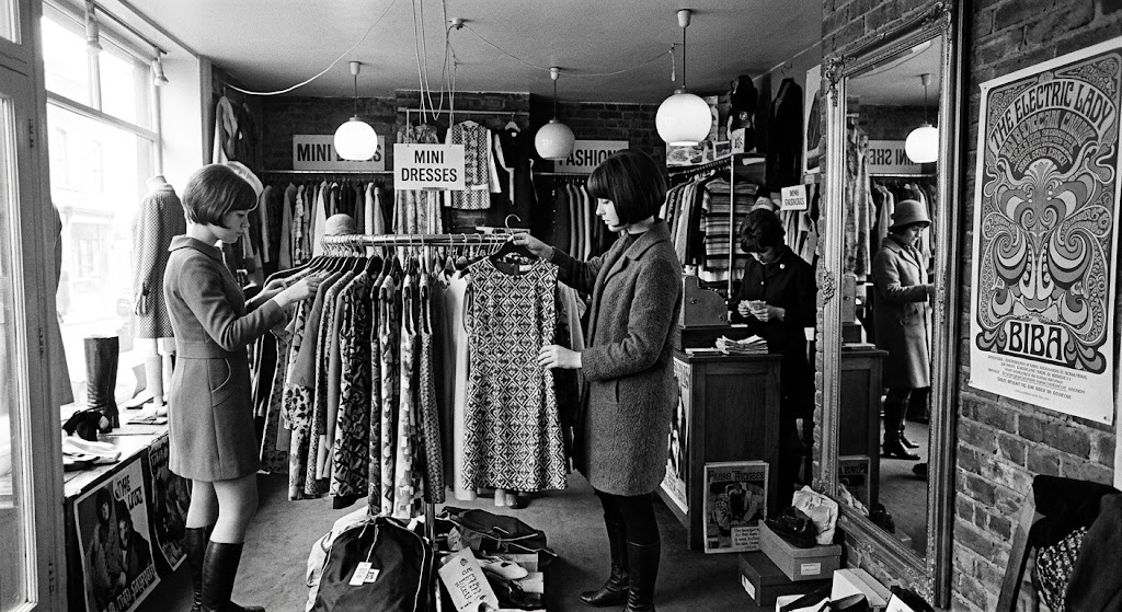 Boutique de moda de los años sesenta con clientas revisando una percha de vestidos